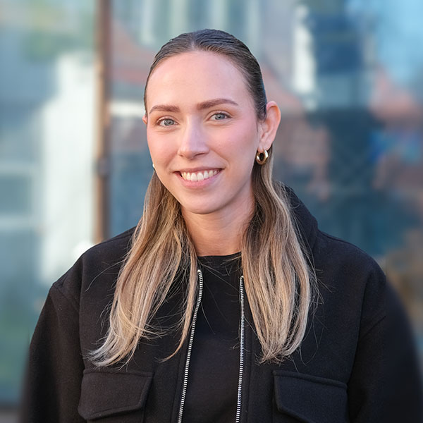 Eine junge Frau steht vor einem verschwommenen Hintergrund und lächelt in die Kamera. Sie hat braun blonde Haare, blaue Augen und trägt eine schwarze Jacke und goldene Ohrringe.