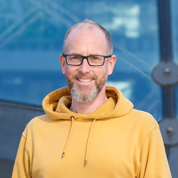 Ein Mann steht vor einem verschwommenen Hintergrund und lächelt in die Kamera. Er hat sehr kurze braune Haare, einen Bart und eine schwarze Brille. Er trägt einen gelben Kapuzenpullover.