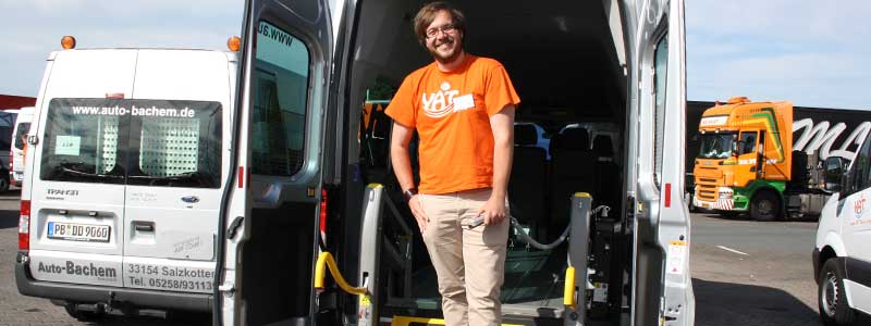 Ein Mann steht lächelnd am offenen Heck eines YAT Transporters mit Rampe, trägt ein orangefarbenes YAT T-Shirt und befindet sich auf einem Parkplatz mit weiteren Fahrzeugen.