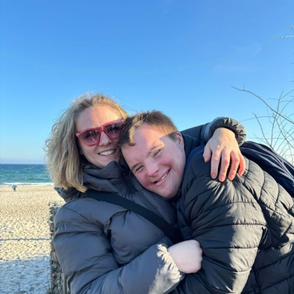 IMG_1731 (1) Eine Frau und ein junger Mann umarmen sich fröhlich und lächeln dabei in die Kamera. Im Hintergrund ist der Strand und das blaue Meer. Der himmel ist blau und wolkenfrei.