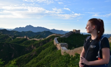 Eine junge Frau mit Rucksack steht seitlich im Bild und blickt über die eindrucksvolle Landschaft mit der Chinesischen Mauer, die sich durch die grünen Berge schlängelt. Der Himmel ist blau mit vereinzelten Wolken.