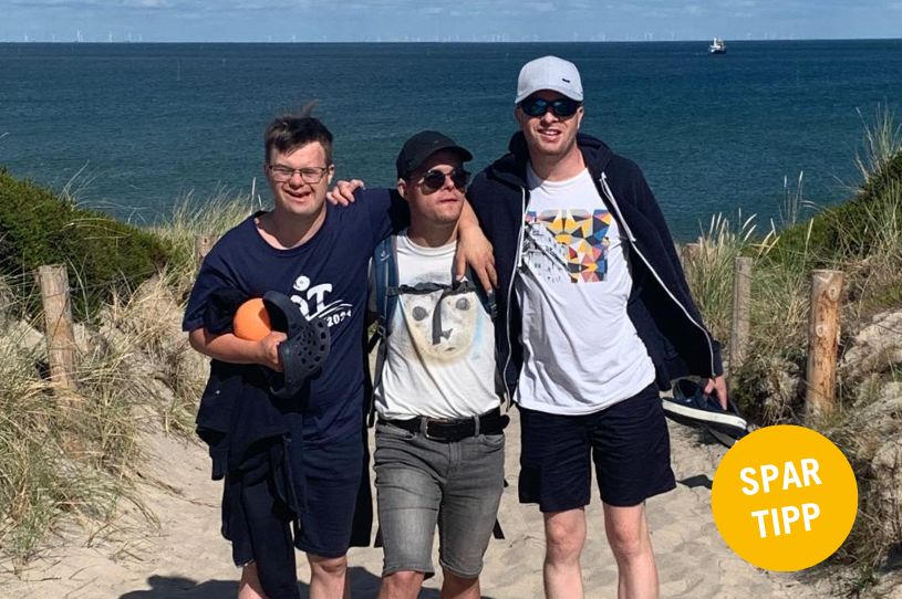 Symbolbild für die YAT-Spartipp Reisen. Darauf sind drei junge Männer mit einer geistigen Behinderung zu sehen. Sie haben sich untergehagt und stehen auf einem Weg zwischen einer Sanddüne. Im Hintergrund ist das Meer zu sehen.