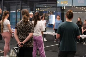 Vier Personen stehen mit dem Rücken zur Kamera, 3 seitlich. Sie hören jemandem zu, der etwas erklärt. Die Gruppe steht auf einem Pedal-Tennis Feld mit einem Netz in der Mitte.