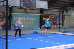 Zwei Personen, eine weiblich, eine männlich stehen in einer Halle auf einem Pedaltennis-Spielfeld. Der Boden ist blau. Sie spielen im Doppel. Sie gucken konzentriert in Richtung der Gegner.