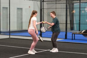 Zwei weibliche Personen in Sportkleidung, die sich einen Tennisball geben. Sie haben beide einen Schläger in der Hand. Sie stehen in einer Halle auf einem Padeltennis Spielfeld.