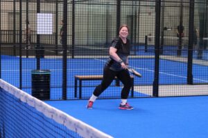Eine Frau läuft über ein Padeltennis-Spielfeld. Sie hält einen Schläger in der Hand. Sie hat Spaß. Der Boden ist blau. Im Hintergrund sieht man ein weiteres Spielfeld.