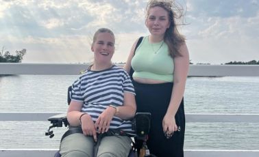 Man sieht eine junge Frau im Rollstuhl und eine stehende junge Frau. Sie gucken beide in die Kamera. Im Hintergrund sieht man das Meer. Es ist extrem windig, sodass die Haare fliegen.