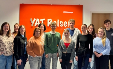 Zwölf Personen stehen vor einer Wand mit einem großen orangenen Schild mit den Buchstaben YAT drauf. Sie gucken lächelnd in die Kamera.