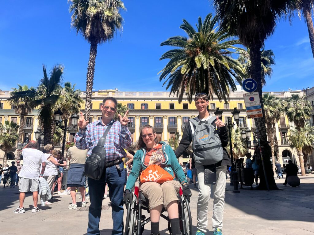 Drei Personen auf einem sonnigen Stadtplatz mit Palmen – einer sitzt im Rollstuhl, alle drei lachen und zeigen das Peace-Zeichen. Gruppenreise mit YAT Reisen.