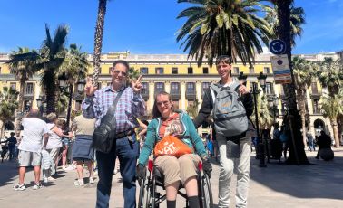 blog-beitragsbild-finanzierung Drei Personen auf einem sonnigen Stadtplatz mit Palmen – einer sitzt im Rollstuhl, alle drei lachen und zeigen das Peace-Zeichen. Gruppenreise mit YAT Reisen.