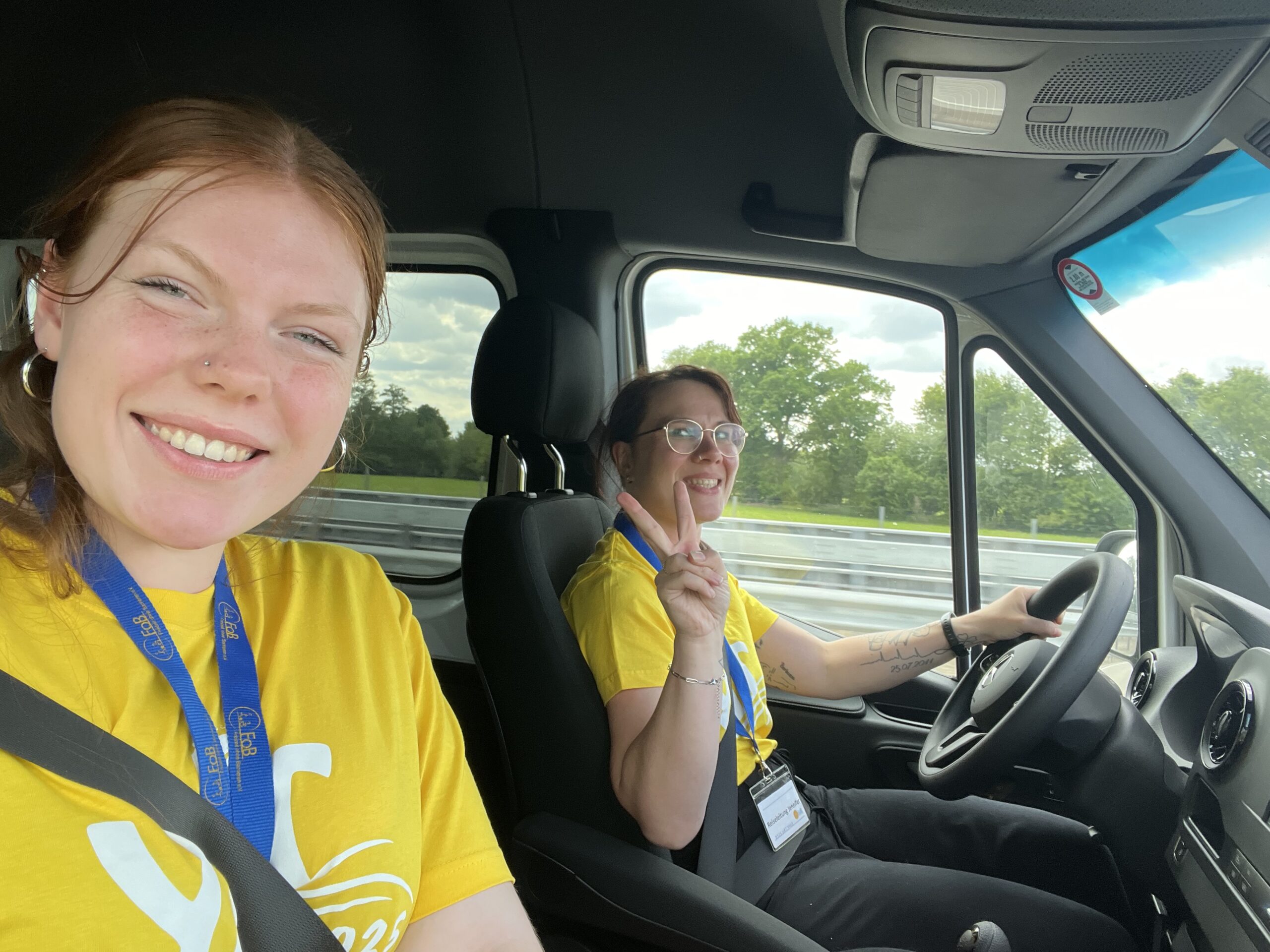 Zwei lächelnde junge Frauen in gelben T-Shirts fahren in einem Transporter auf der Autobahn – die Fahrerin zeigt ein Peace-Zeichen.