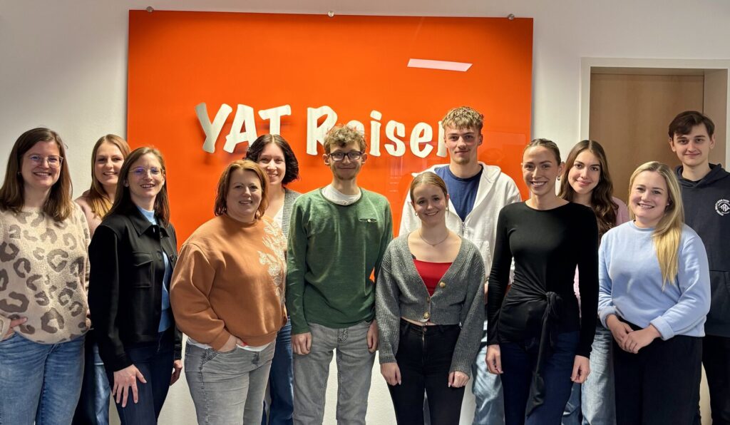 Zwölf Personen stehen vor einer Wand mit einem großen orangenen Schild mit den Buchstaben YAT drauf. Sie gucken lächelnd in die Kamera.
