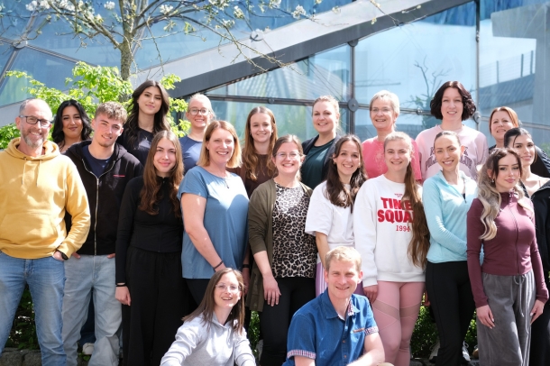 team-gruppenfoto Gruppenfoto eines Teams von etwa 18 Personen vor einer modernen Glasfassade, aufgestellt in mehreren Reihen im Freien bei Sonnenschein, im Hintergrund ein blühender Baum.