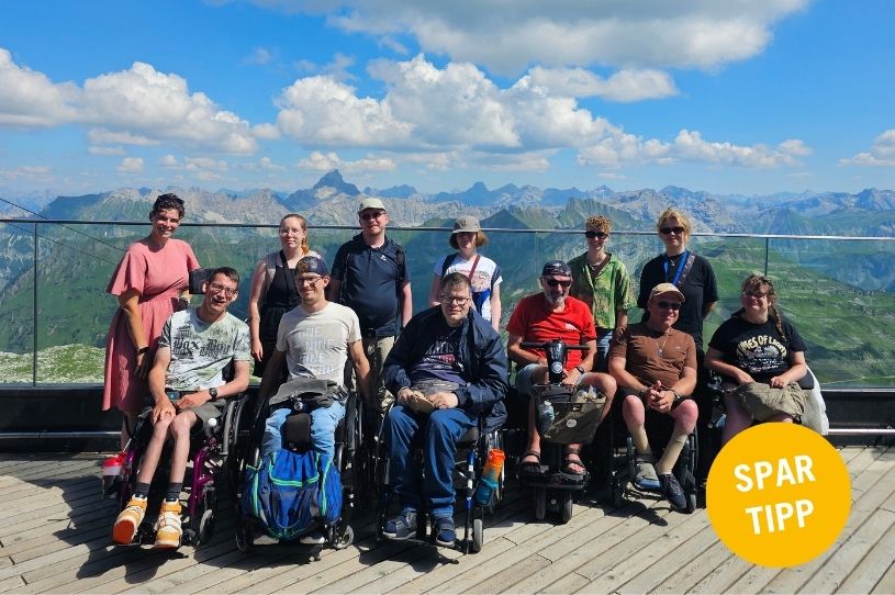Eine Gruppe posiert für ein Foto. In der vorderen Reihe sind Personen zu sehen, die im Rollstuhl sitzen. Die Personen in der hinteren Reihe stehen. Im Hintergrund ist eine Berglandschaft zu sehen.